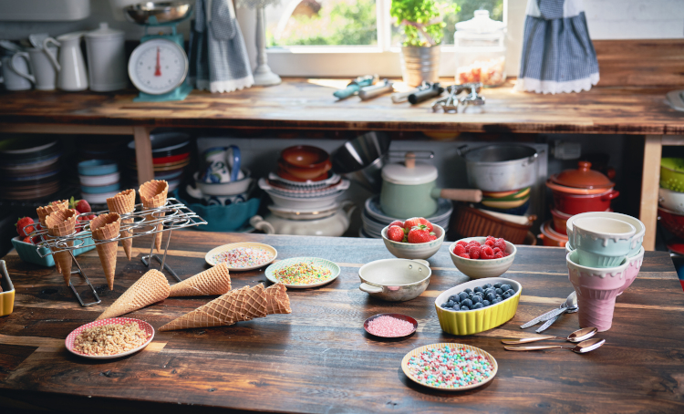 A table with waffle cones, sprinkles, freeze dried strawberries, crushed nuts, berries, spoons, ice cream cone holder.