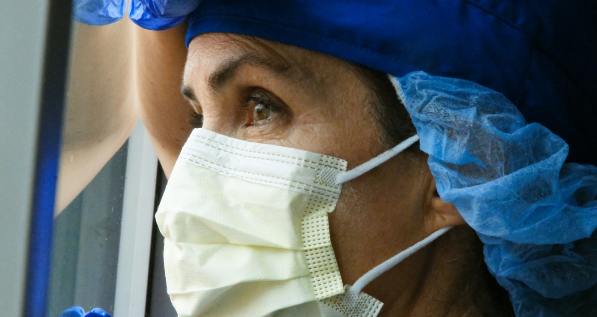 A health worker in PPE resting her hand and head against a window and looking outside.