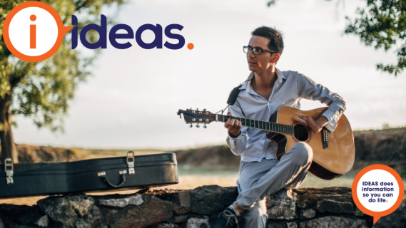 A young man with disability plays a guitar. He sits outside on a stone wall, next to him is the guitar case.